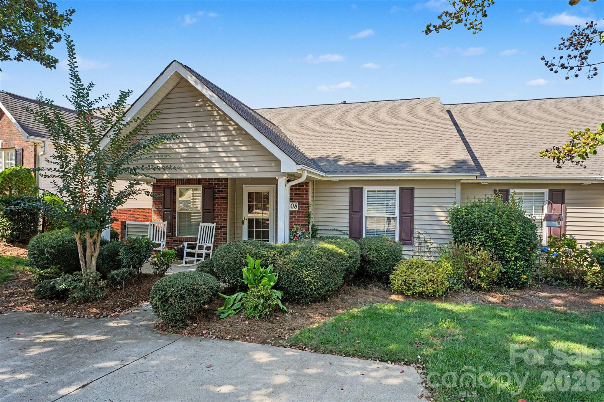 Single-story gray-sided home with a brick entry, landscaped shrubs, and two white rocking chairs on the porch.