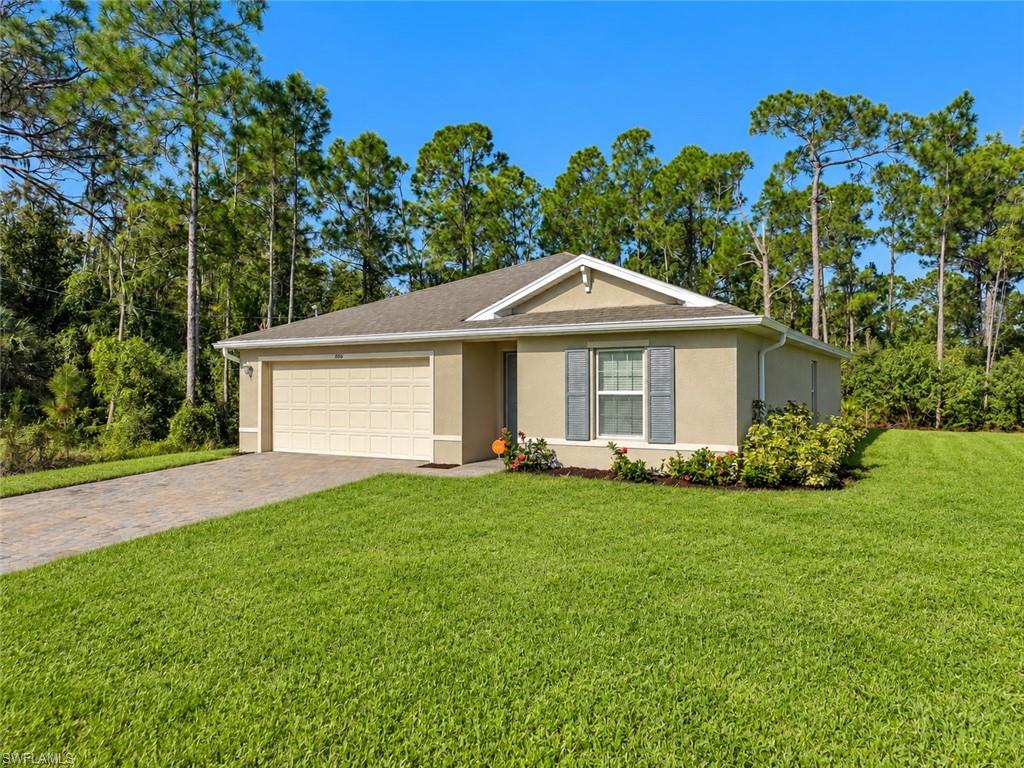 Beige single-story home with a two-car garage, paved driveway, and green lawn framed by tall pines.
