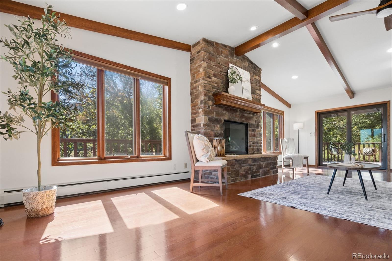 Living room with a stone fireplace, exposed wood beams, and large windows overlooking trees.