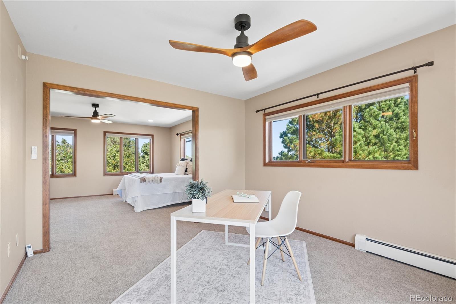 Bedroom with an adjoining work area, wood-trimmed windows, leafy views, and a ceiling fan.