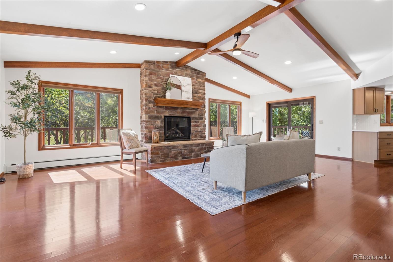 Living room with a stone fireplace, vaulted wood-beam ceiling, hardwood floors, and large windows.
