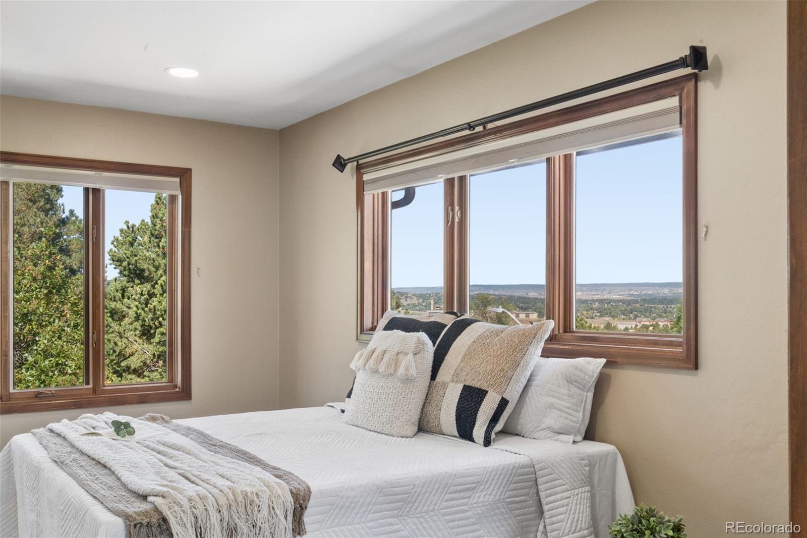 Staged bedroom with a white bed, wood-trimmed windows, and a view of trees and distant hills.