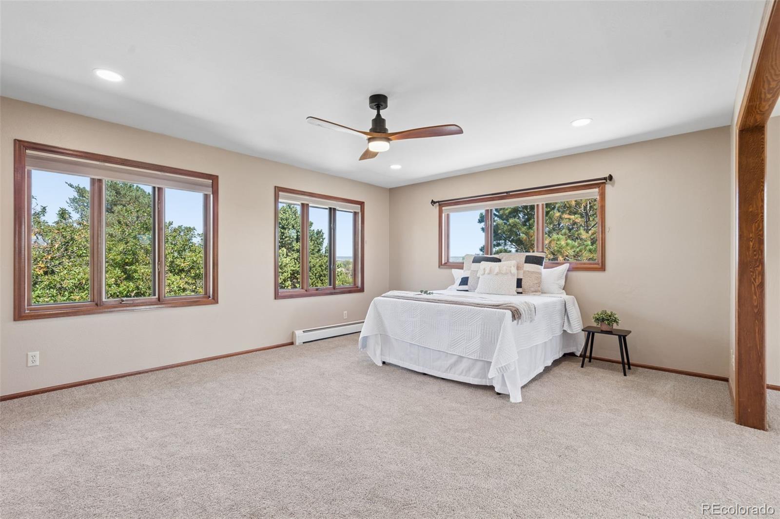 Staged bedroom with a white bed, beige carpet, wood-trimmed windows, and a ceiling fan.