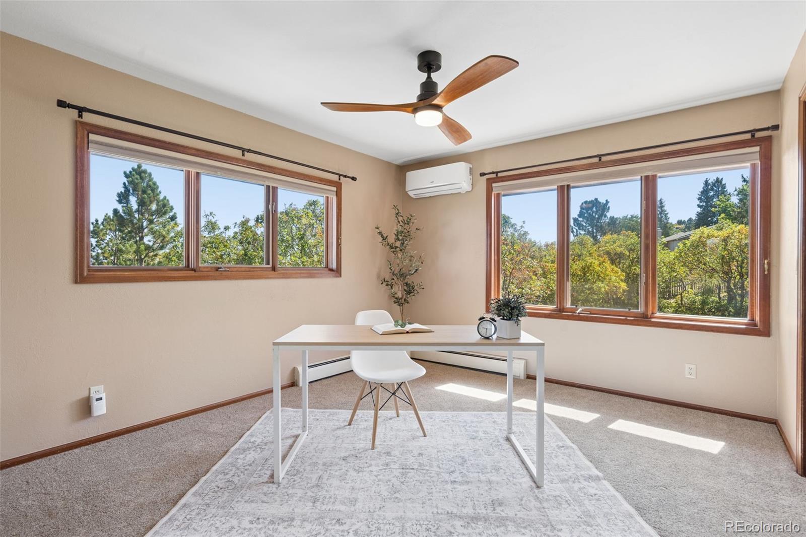 Staged home office with a white desk, wood-trimmed windows, and leafy views.