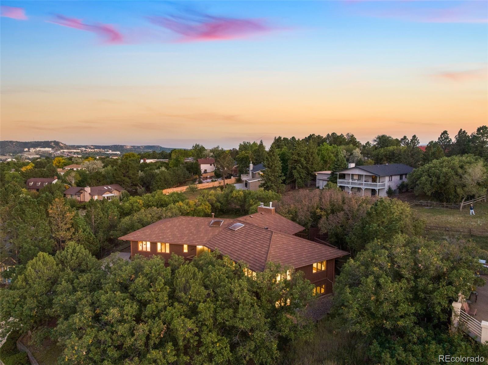 Aerial view of a red tile-roofed home among trees at dusk, with neighboring homes and a colorful sunset sky.