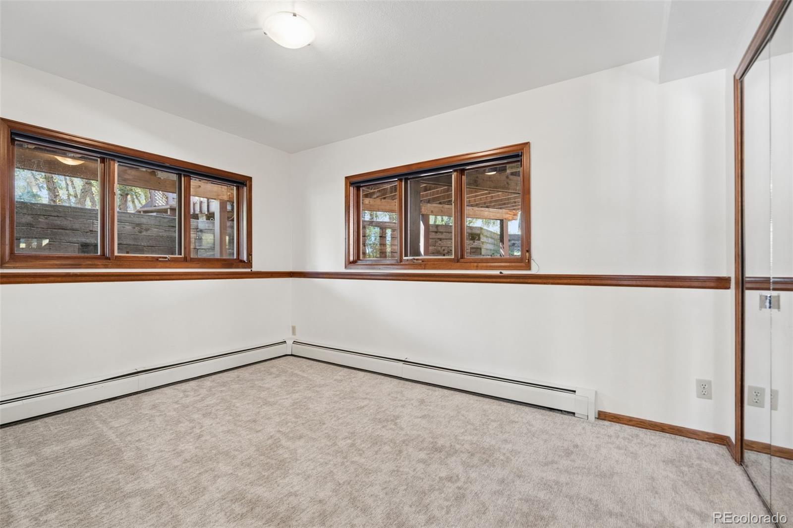 Empty carpeted room with wood-trimmed windows, mirrored closet door, and baseboard heating.
