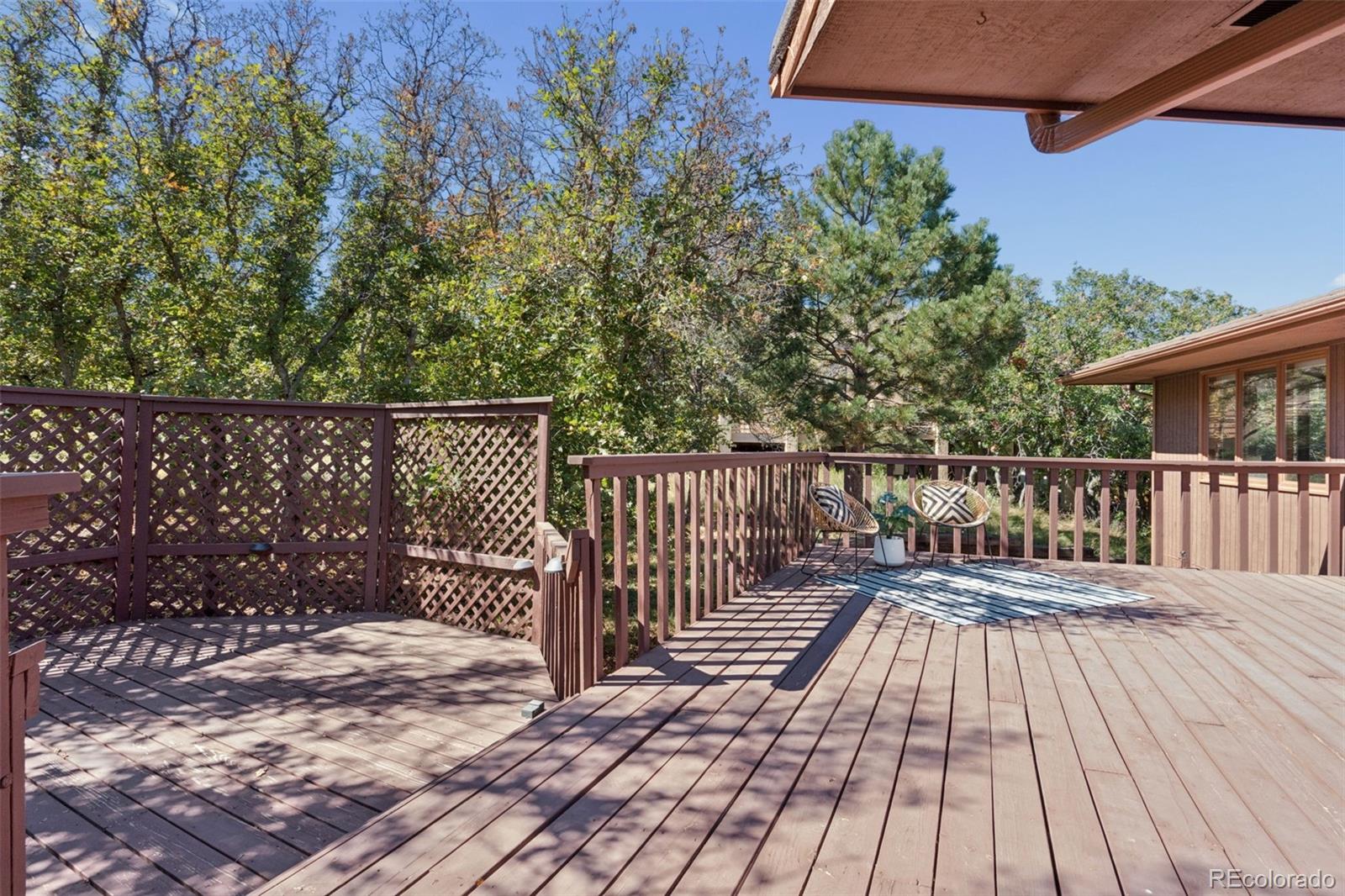 Wooden deck with lattice fencing, two patterned chairs, and mature trees.