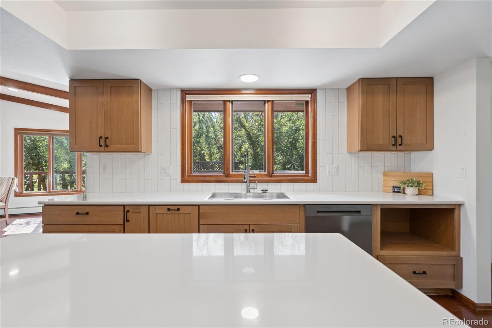 Kitchen with warm wood cabinets, white counters, and a sink beneath a large tree-facing window.