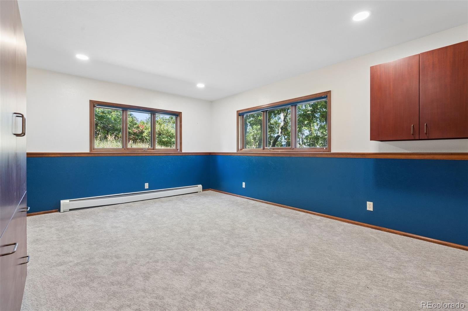 Empty carpeted room with blue walls, built-in cabinets, two windows and a baseboard heater.