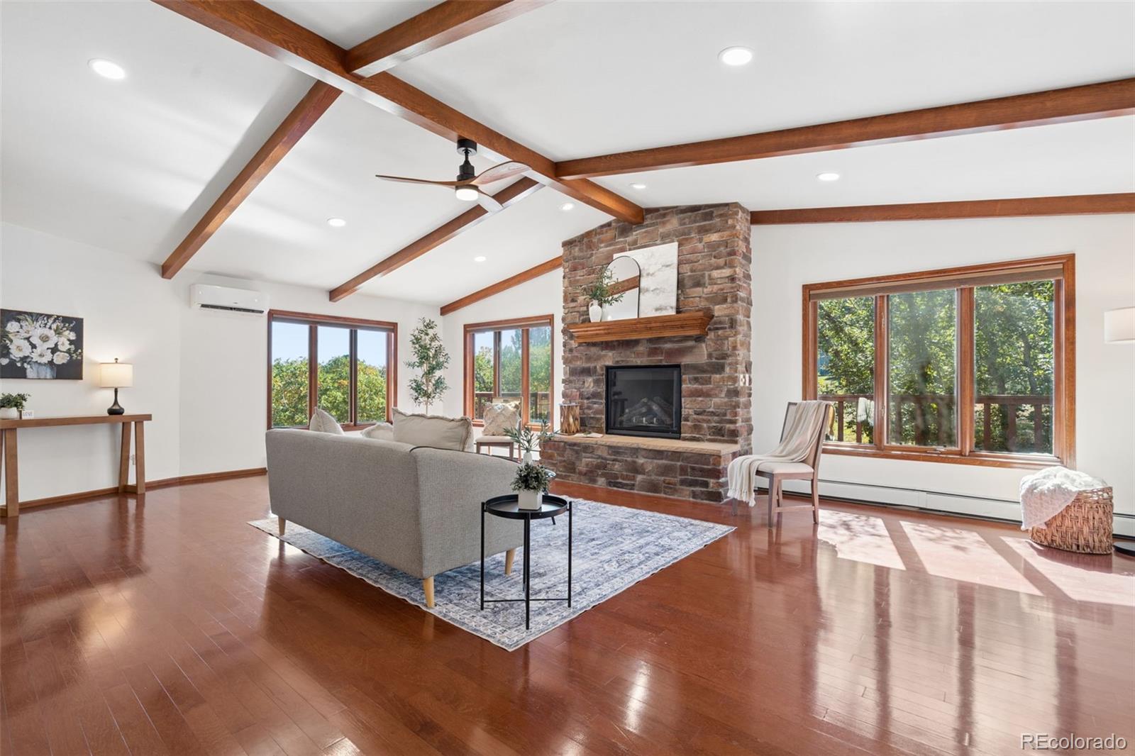 Living room with exposed wood ceiling beams, a stone fireplace, and large windows overlooking trees.