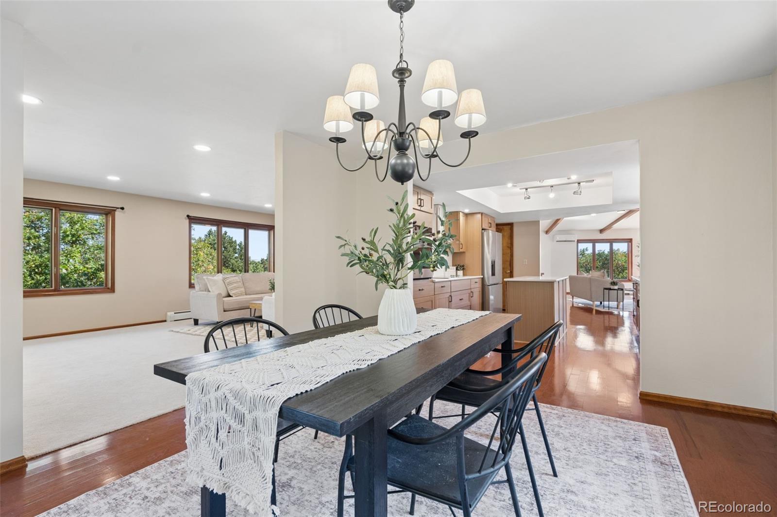 Dining room with a long dark table and chandelier, opening to a living area and kitchen.