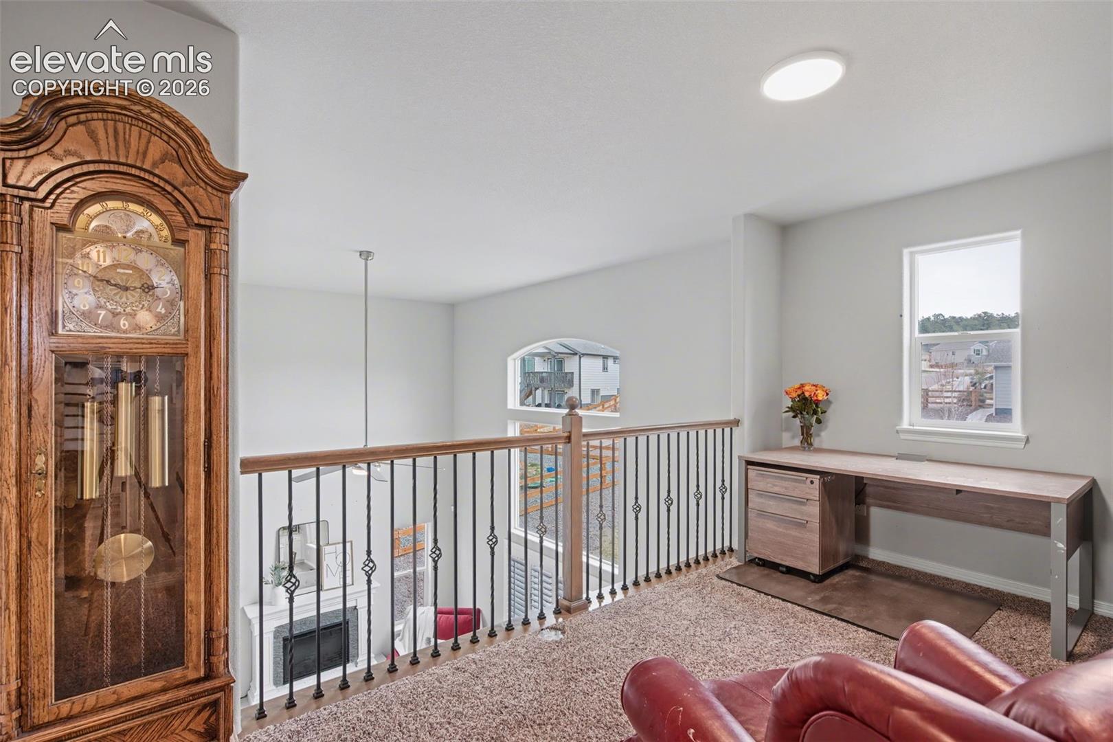 Upstairs loft overlooks a living room, with a wood-and-metal railing, desk, carpet, and grandfather clock.