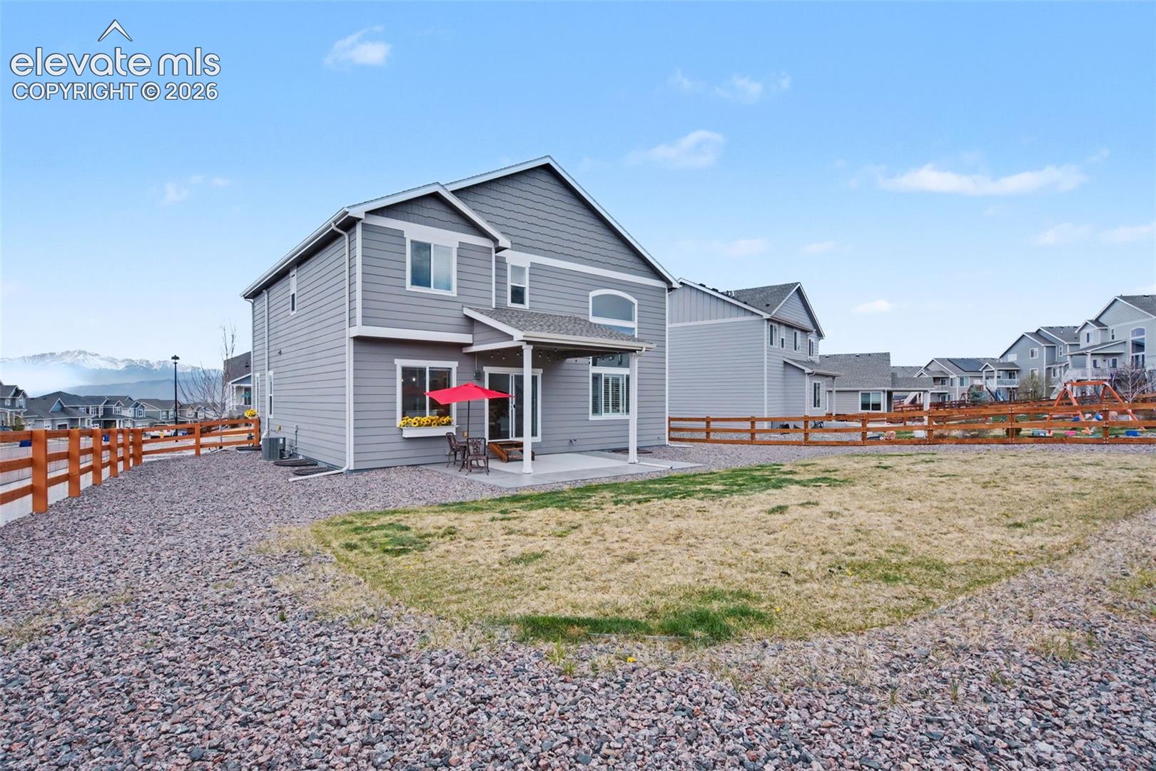 Gray two-story home with a covered patio and red umbrella, fenced yard, and distant snow-dusted mountains.