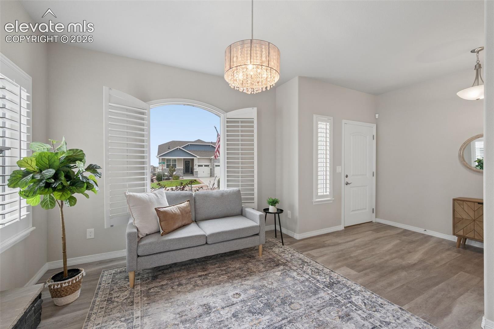 Staged living room with a gray sofa, crystal chandelier, arched shuttered window, and patterned rug.