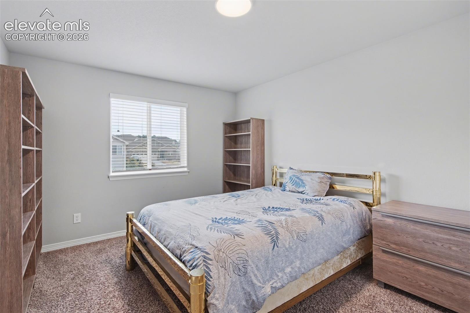 Furnished bedroom with a brass bed, leafy blue bedding, wooden bookcases, and a window with white blinds.