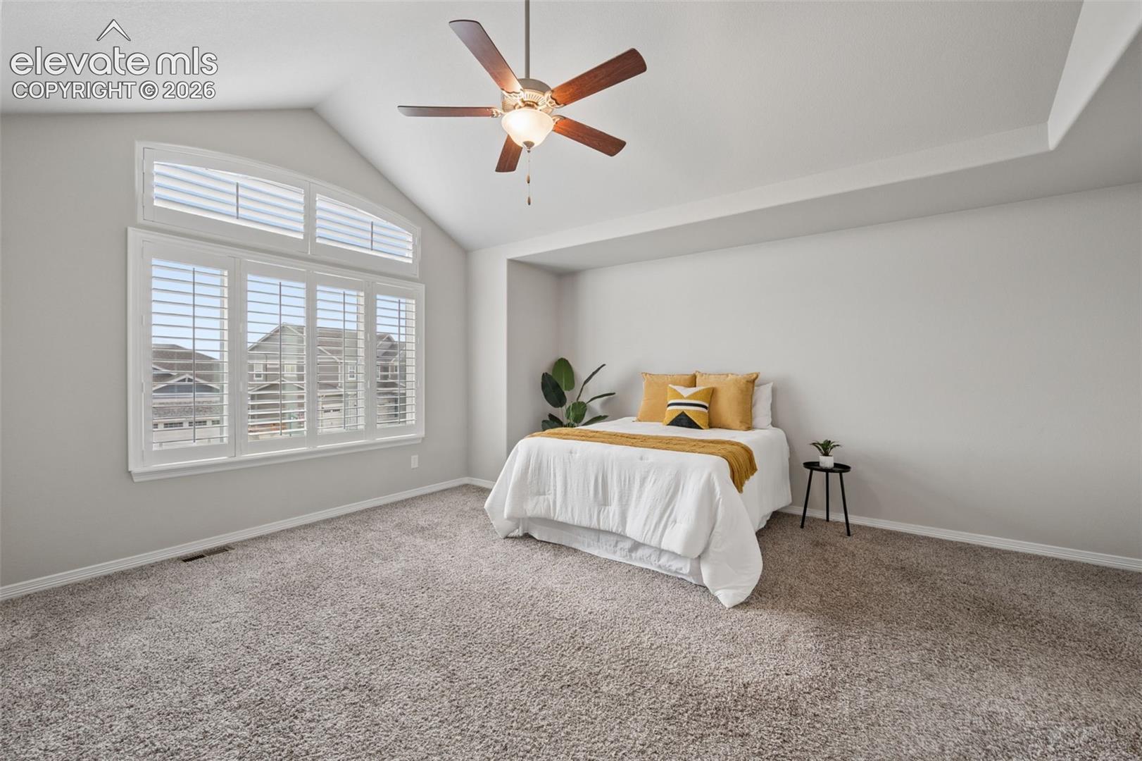 Staged bedroom with a mustard-accented bed, vaulted ceiling, and large plantation-shuttered windows.