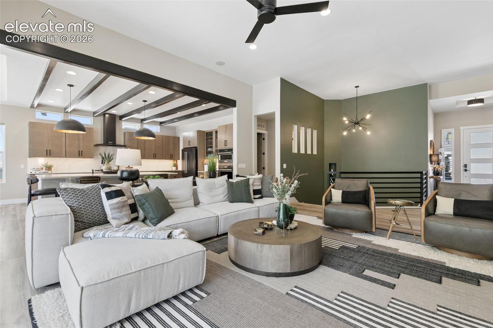 Open-plan living room with a white sectional, gray chairs, and kitchen beneath exposed ceiling beams.