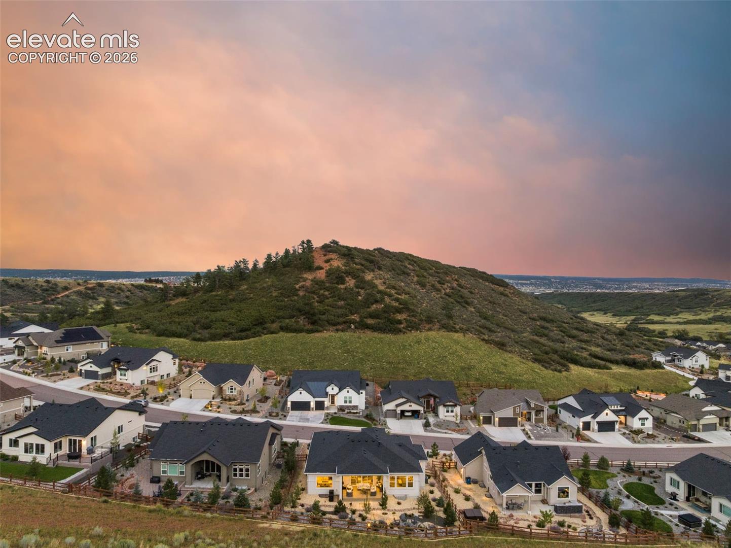 Aerial neighborhood below a scrub-covered hill under a pink and blue dusk sky.