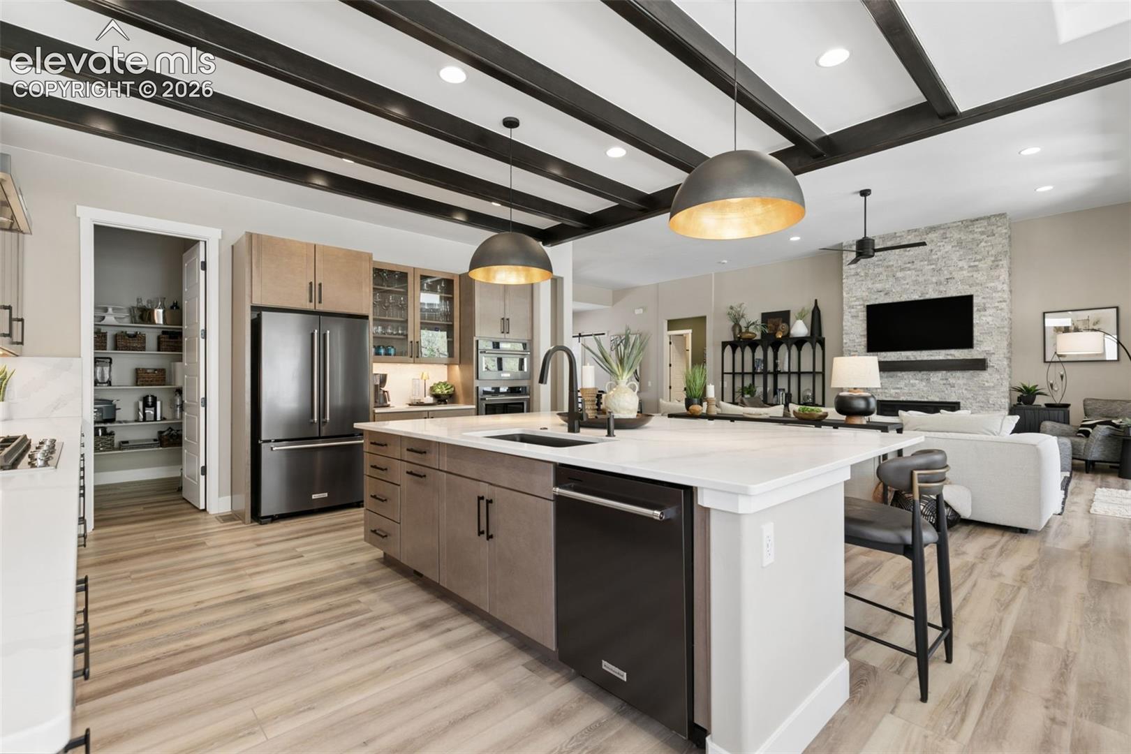 Open-plan kitchen with a large white island, dark ceiling beams, and adjoining living room with stone fireplace.