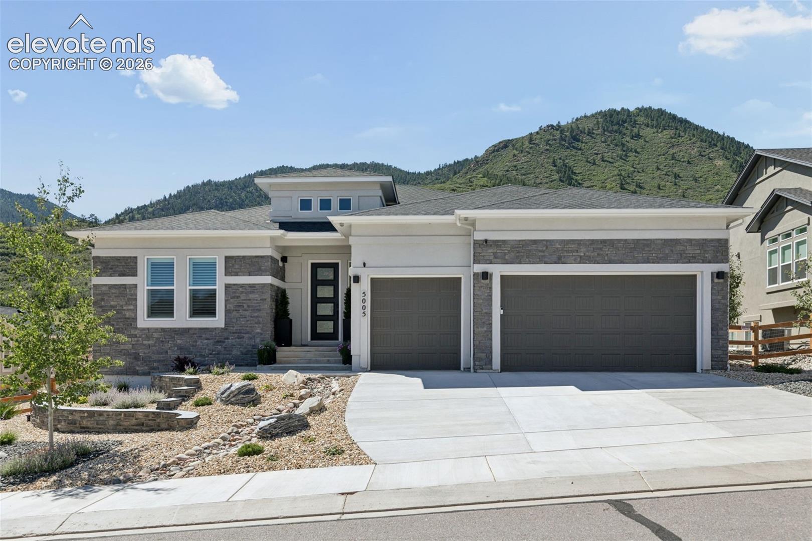 Modern stone-and-siding home with a two-car garage, rock garden, and wooded mountain backdrop.