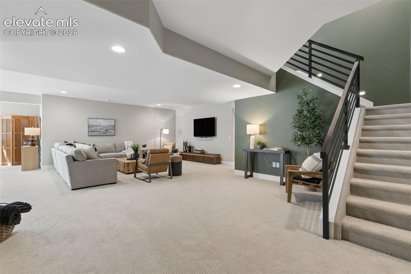 Carpeted living room with a gray sectional, wall-mounted TV, and staircase with dark railing.