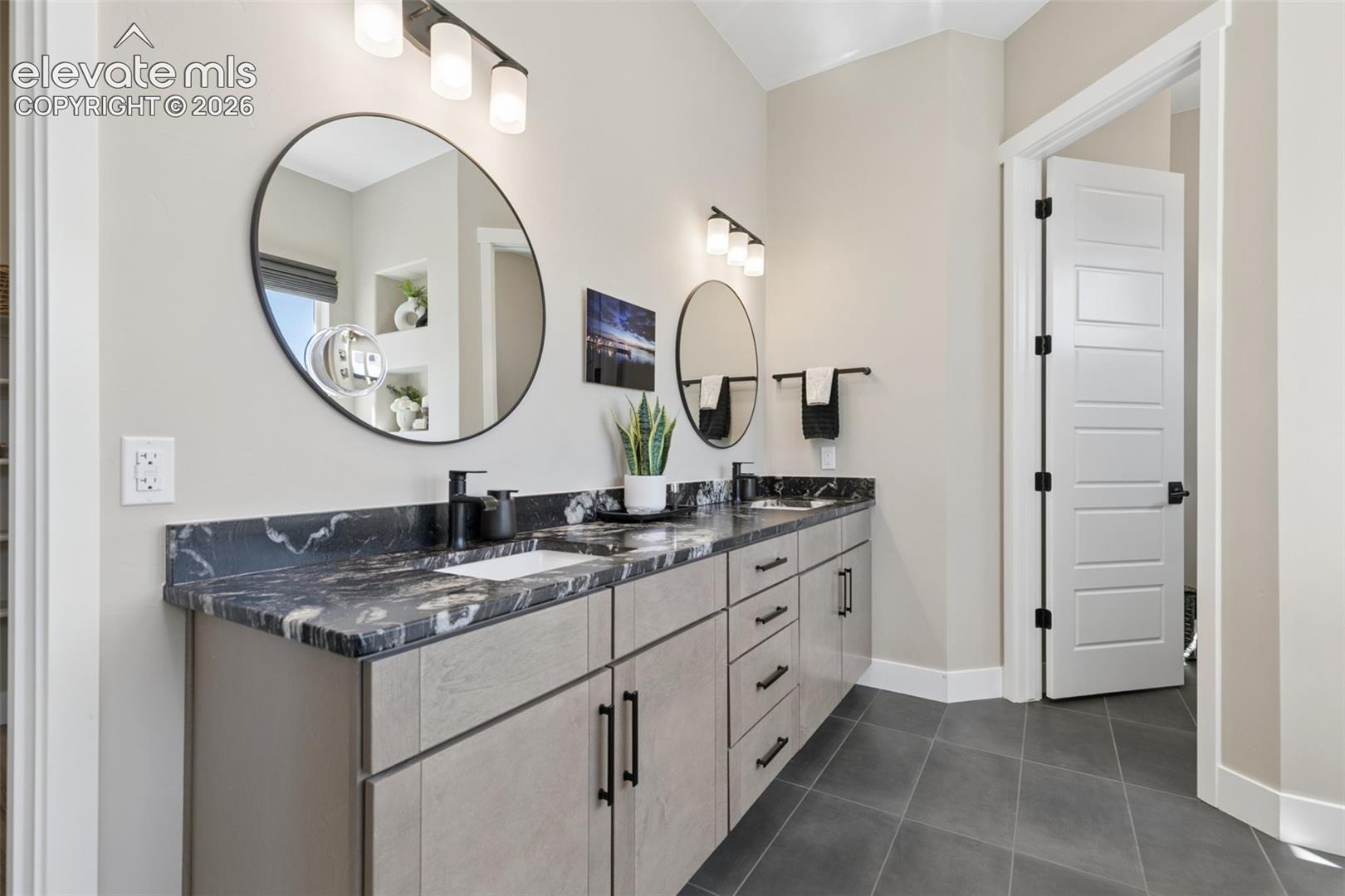 Bathroom with a double vanity, veined stone countertop, round mirrors, and dark tile floor.