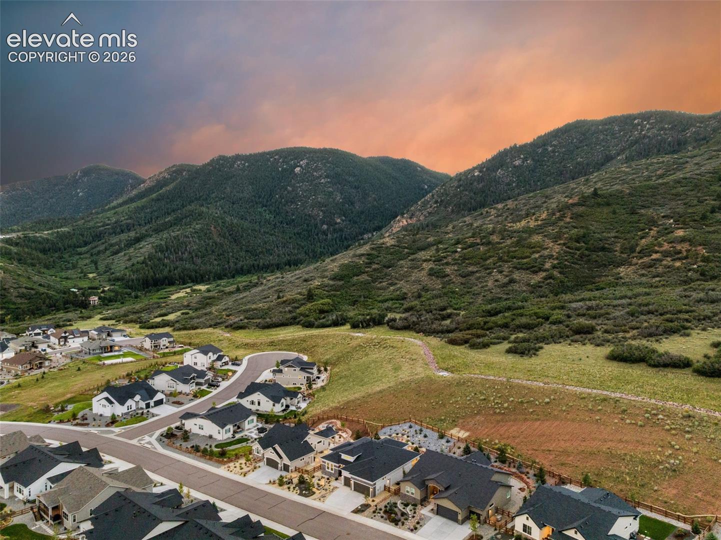 Aerial view of homes beside forested mountains beneath a vivid orange sunset sky.