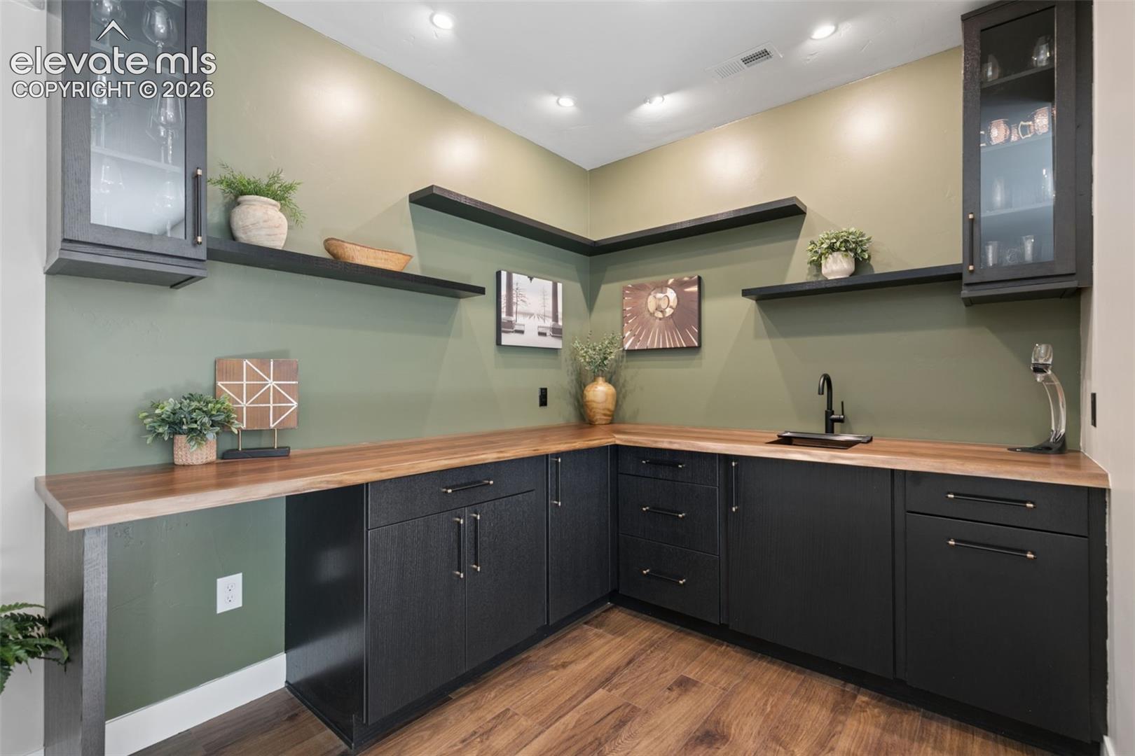 Wet bar with dark cabinets, wood countertop, floating shelves, and a black sink faucet.