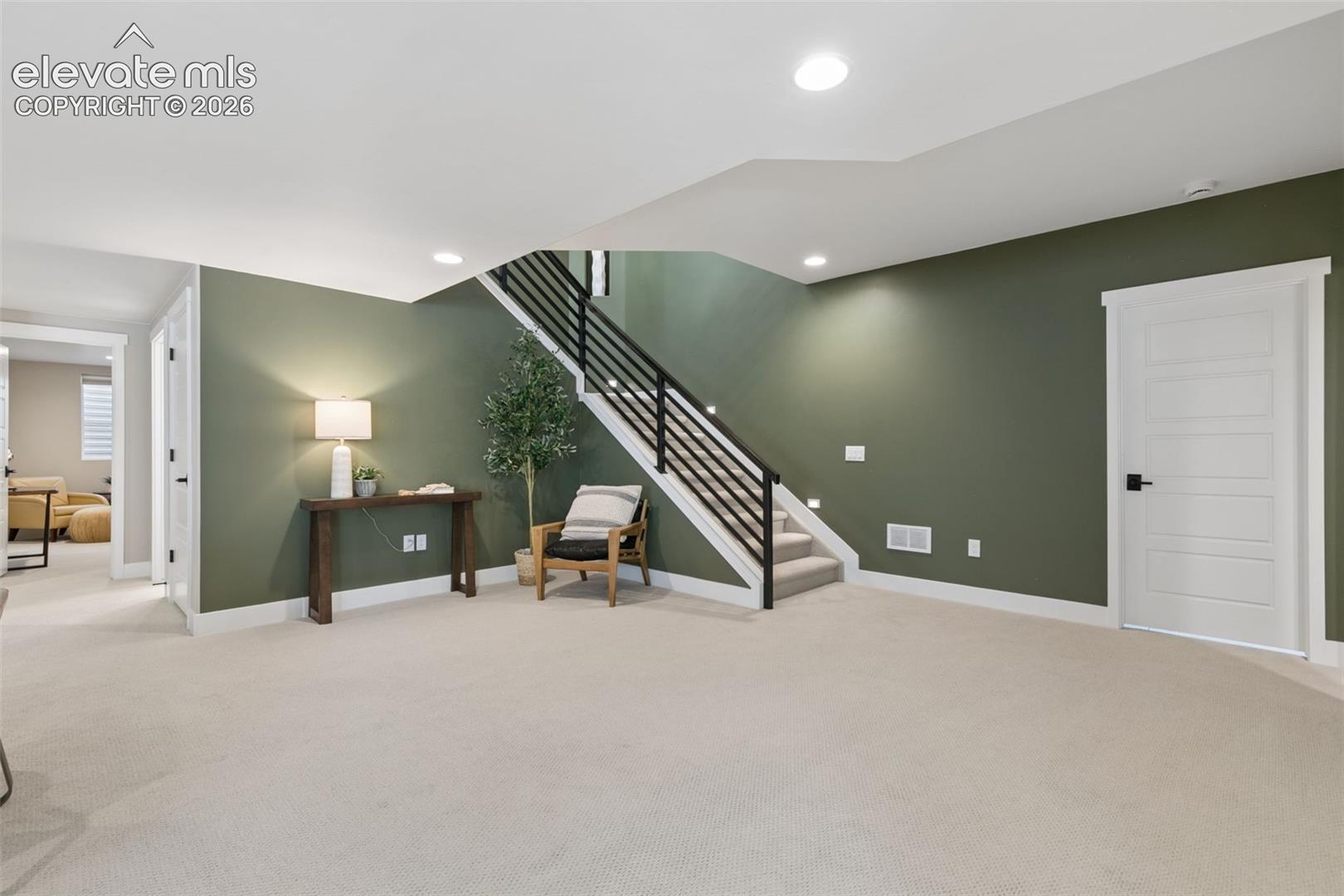Carpeted lower-level living area with an olive-green wall and black metal-railed staircase.