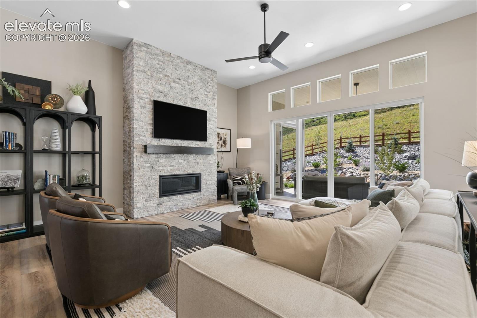 Living room with a stone fireplace, cream sectional, and wide windows overlooking a landscaped hillside.