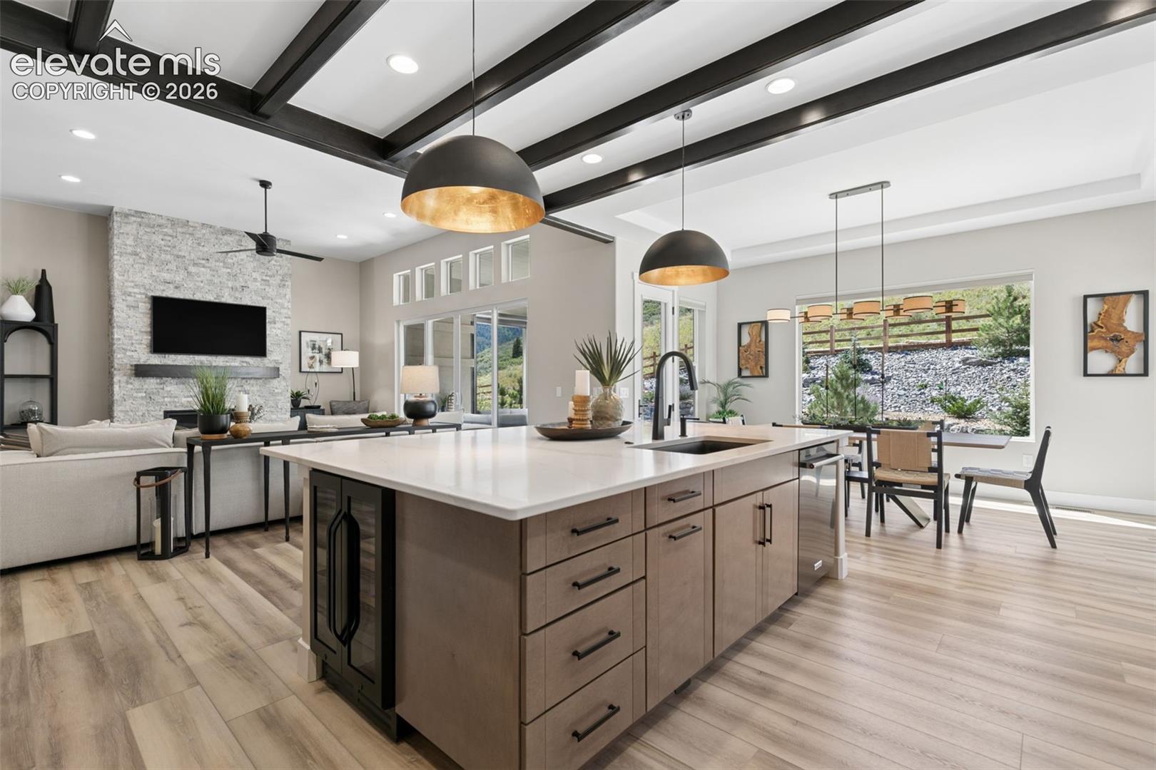 Open-plan kitchen with a large island, dark ceiling beams, and a stone fireplace beside the living area.