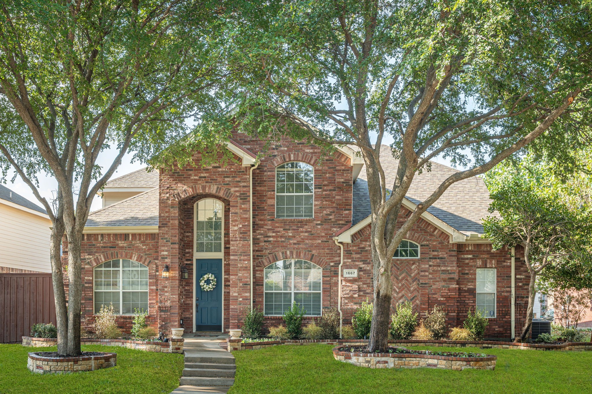 Two-story red brick house with a blue front door framed by mature trees and landscaped gardens.