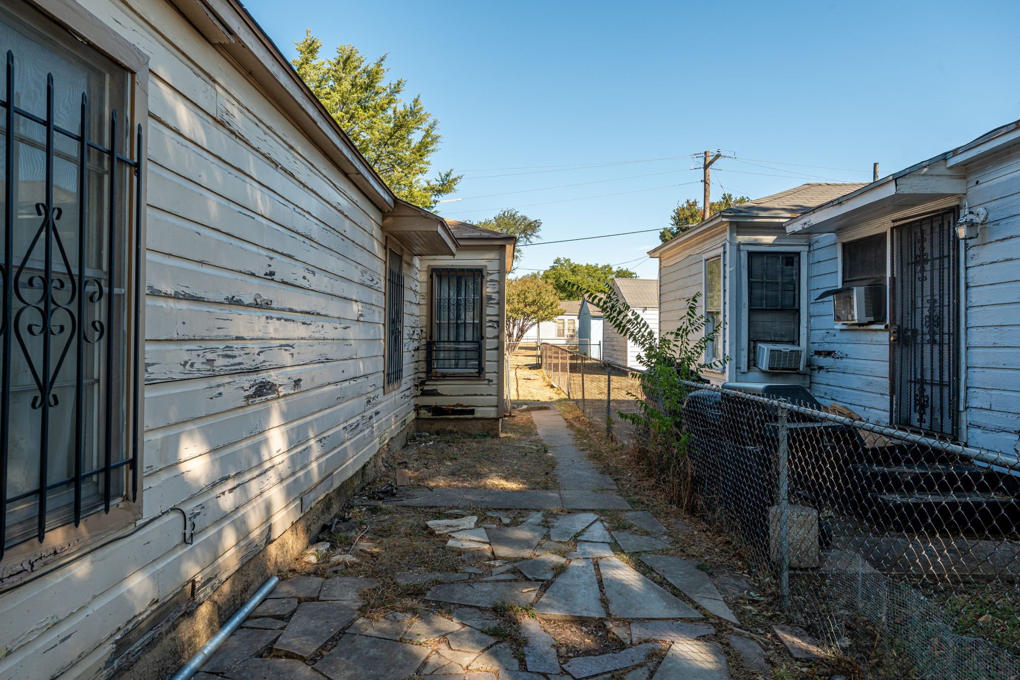 Narrow side yard between weathered houses, with a broken stone path and chain-link fence.