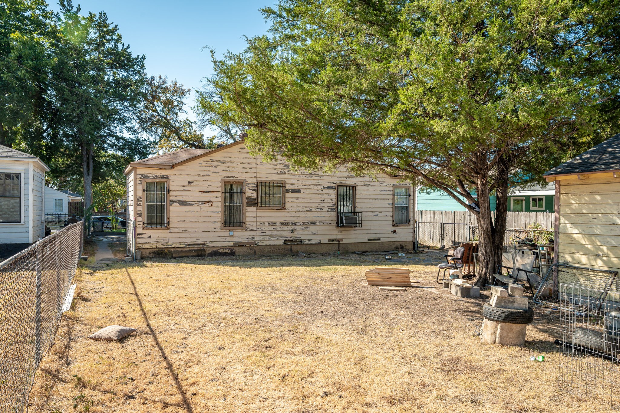 Weathered single-story house with barred windows behind a dry, chain-link-fenced yard and large tree.