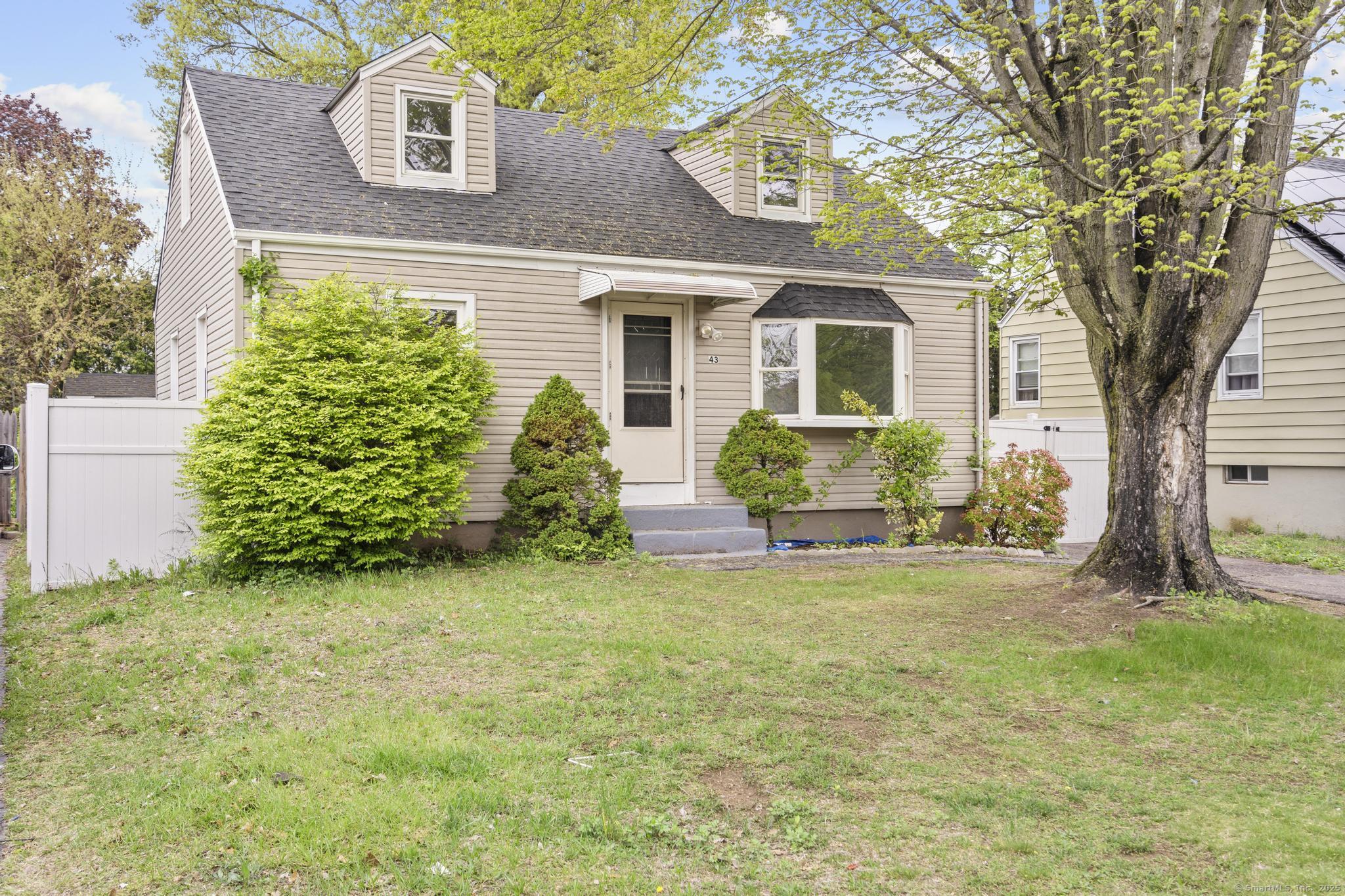 Beige two-story house with dormer windows, a bay window, and a large tree in the front yard.