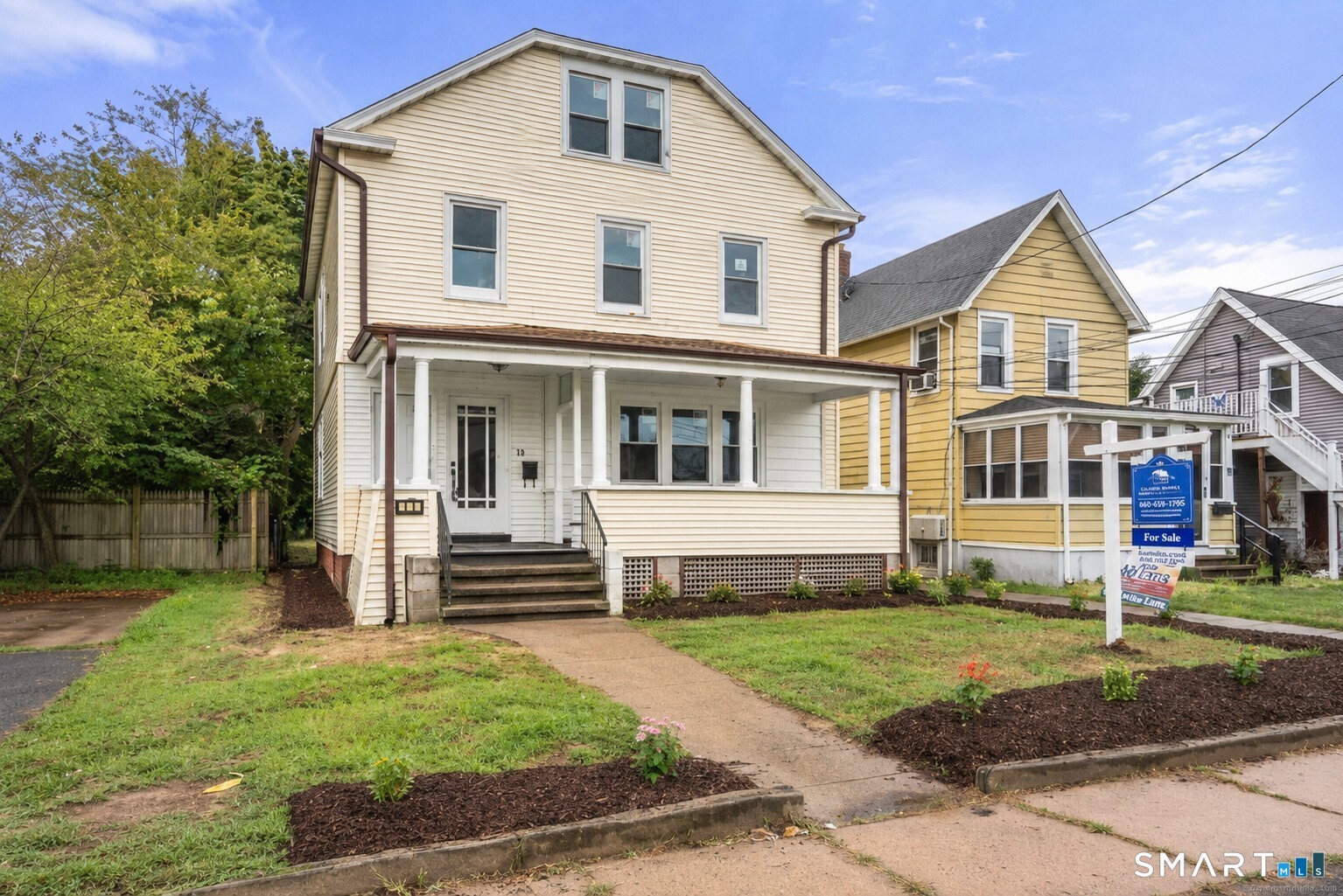 Cream-sided multi-story house with a covered porch, front steps, and a for-sale sign.