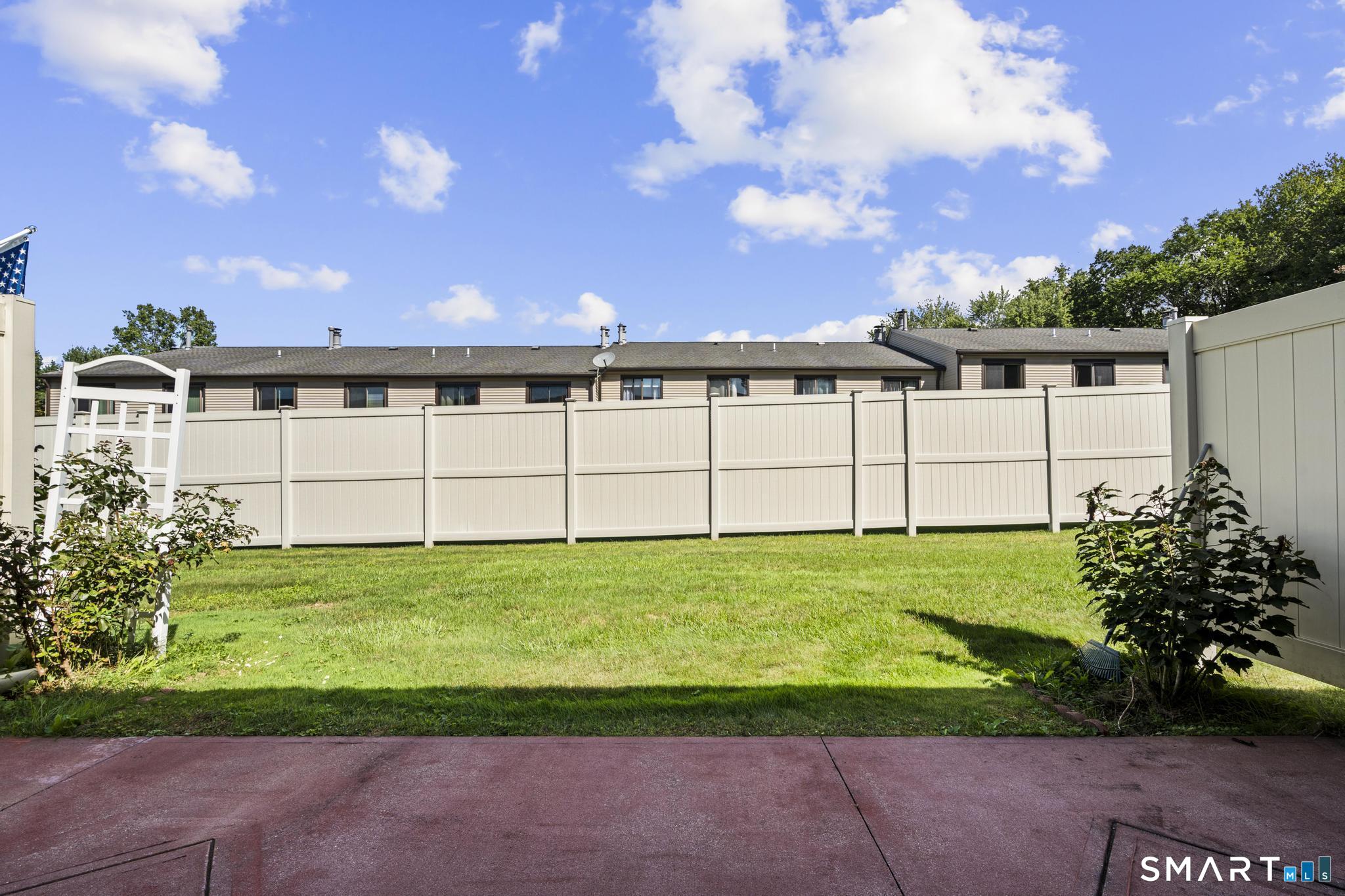 Fenced backyard with lawn, patio, and a white garden arbor behind a condominium.