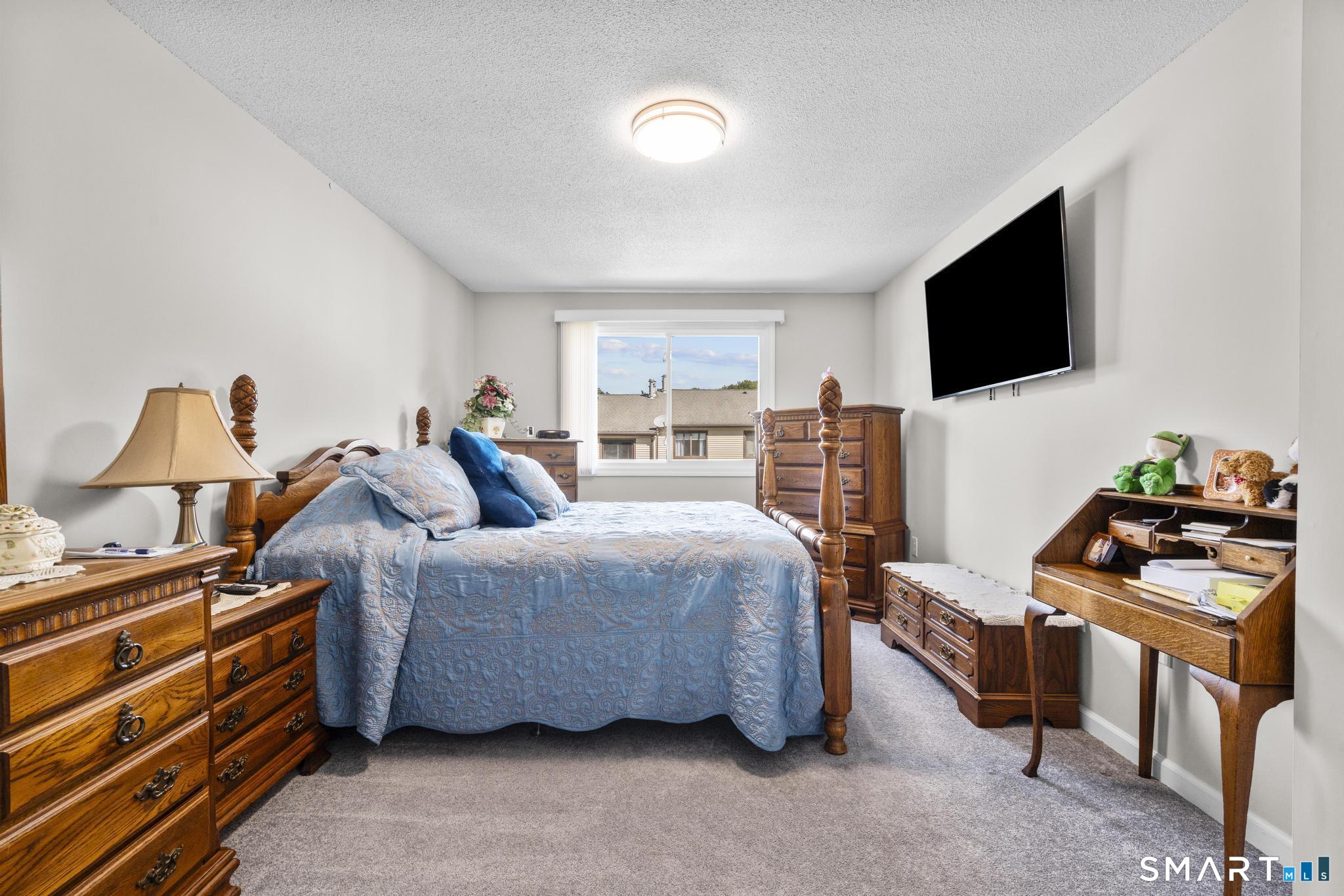 Bedroom with a blue quilted bed, traditional wooden furniture, and a window overlooking neighboring homes.