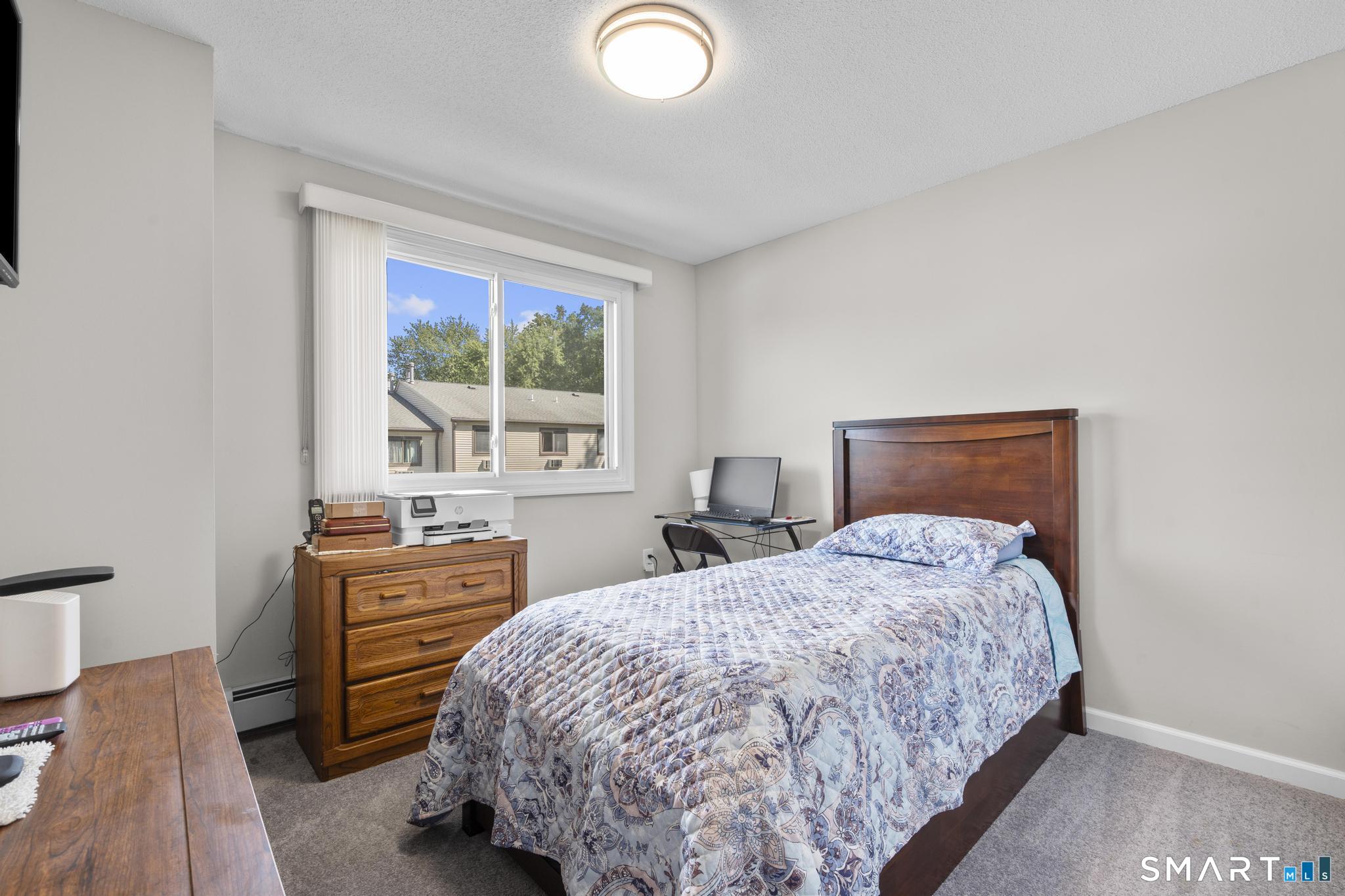 Bedroom with a patterned bedspread, wooden headboard, and window overlooking neighboring homes.