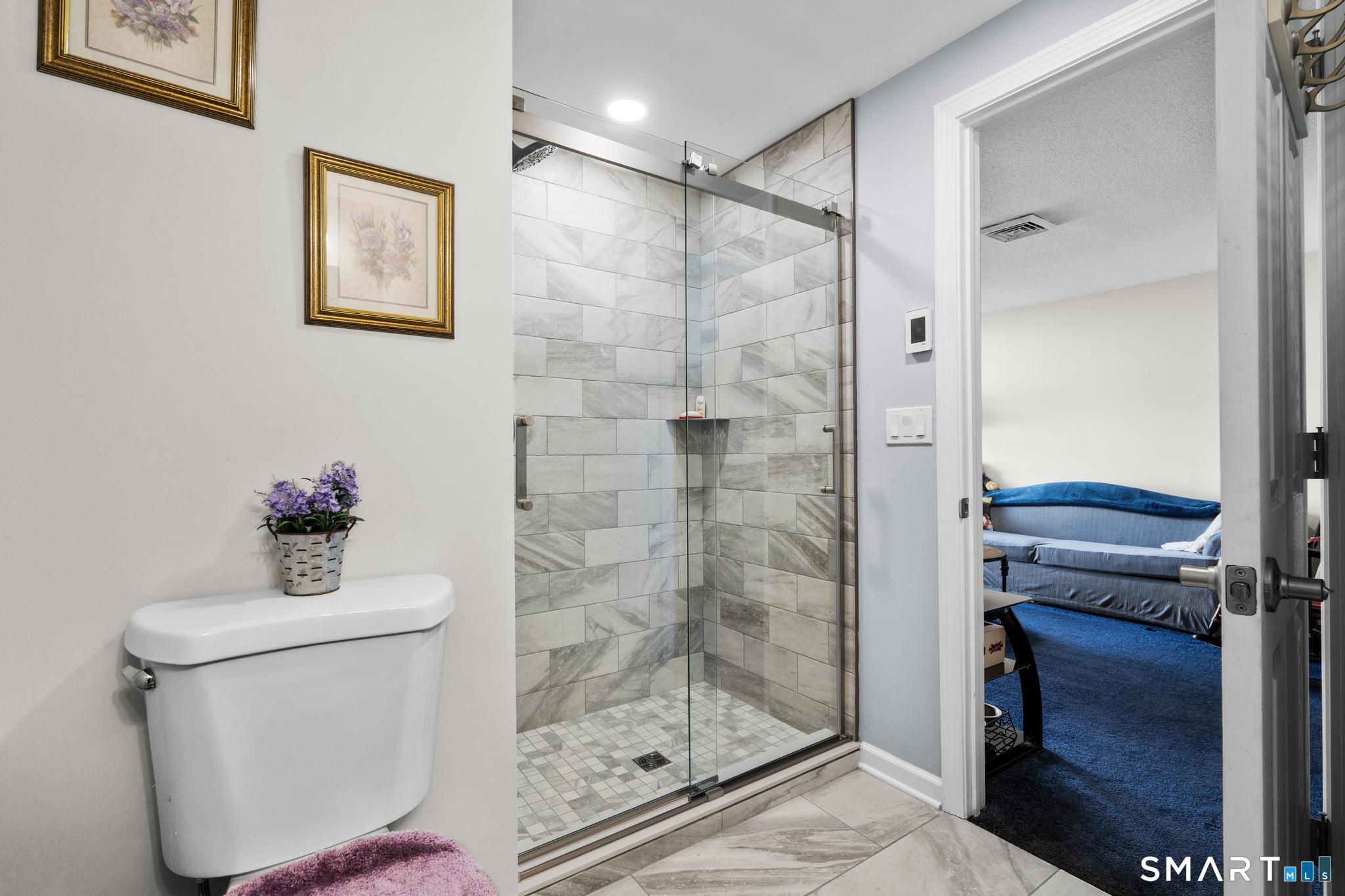 Bathroom with a glass-enclosed marble-look tile shower beside an open doorway to a blue-carpeted room.