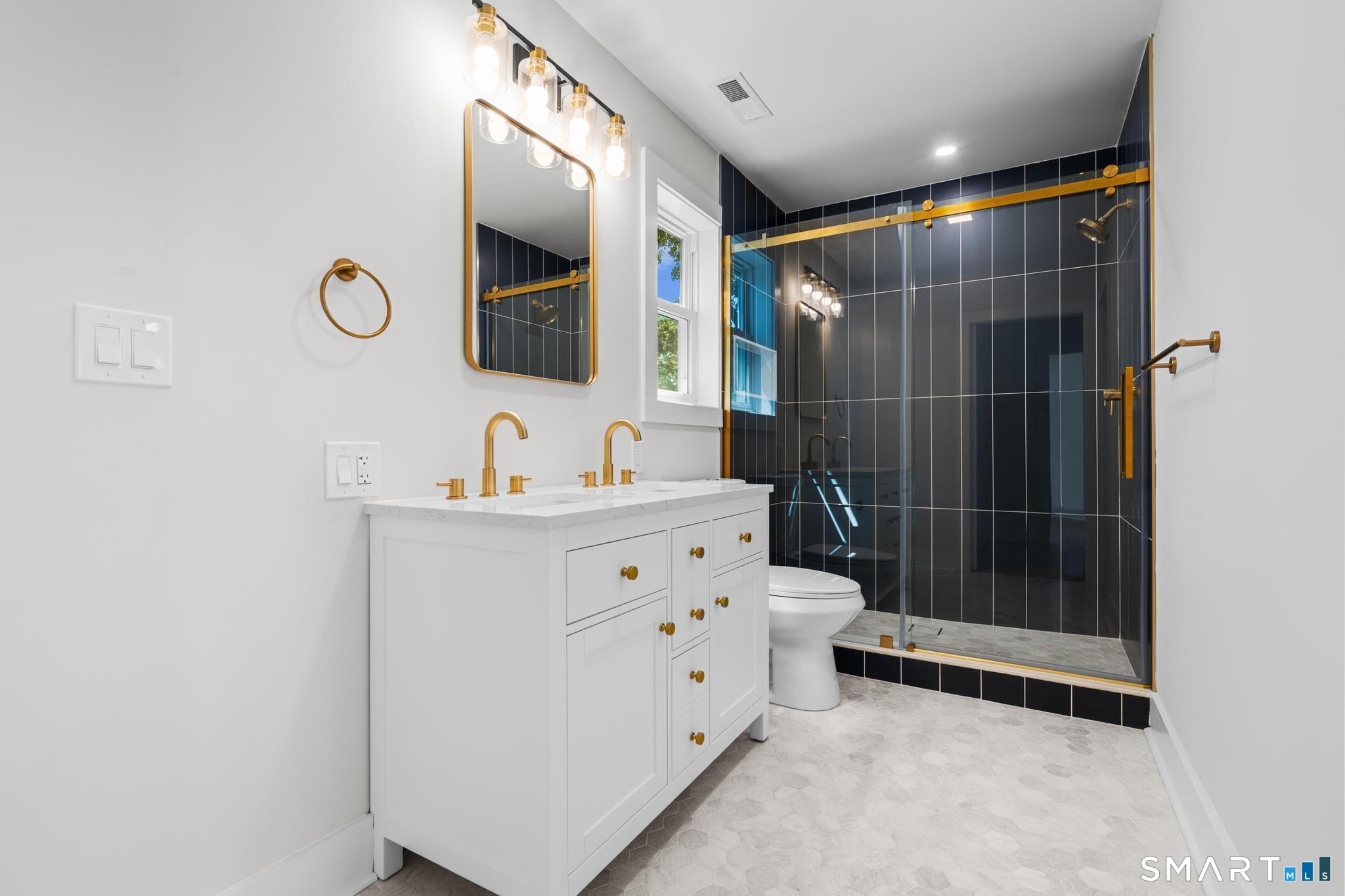 Bathroom with a white double-sink vanity, gold fixtures, and a dark tiled glass shower.