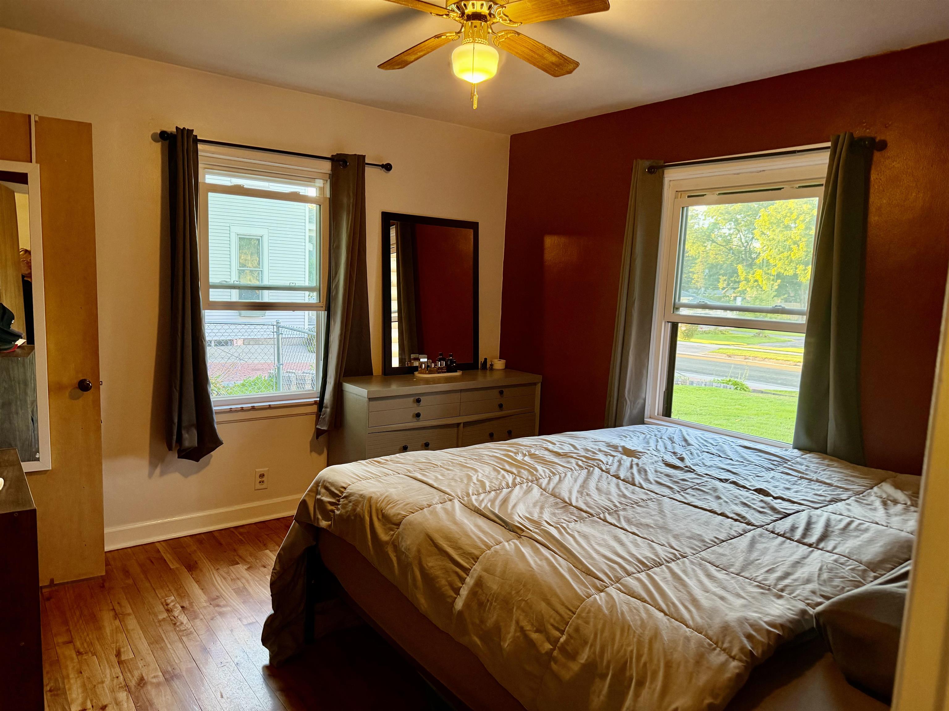 Bedroom with a large bed, hardwood floors, and two windows overlooking the street and yard.