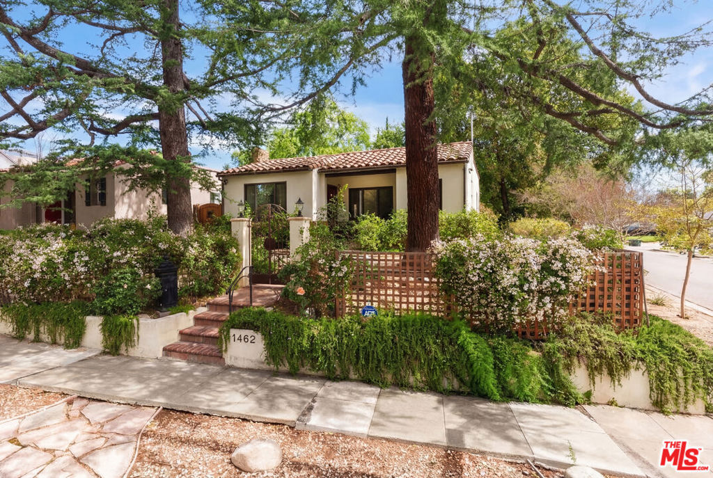 Single-story stucco home with a tile roof, mature trees, flowering shrubs, and a gated entry.