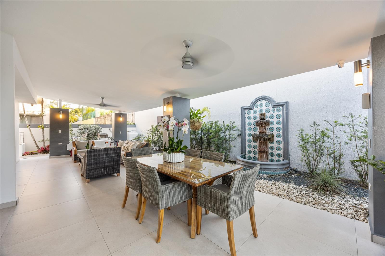 Covered patio with a glass-topped dining table, woven chairs, and a tiled fountain.
