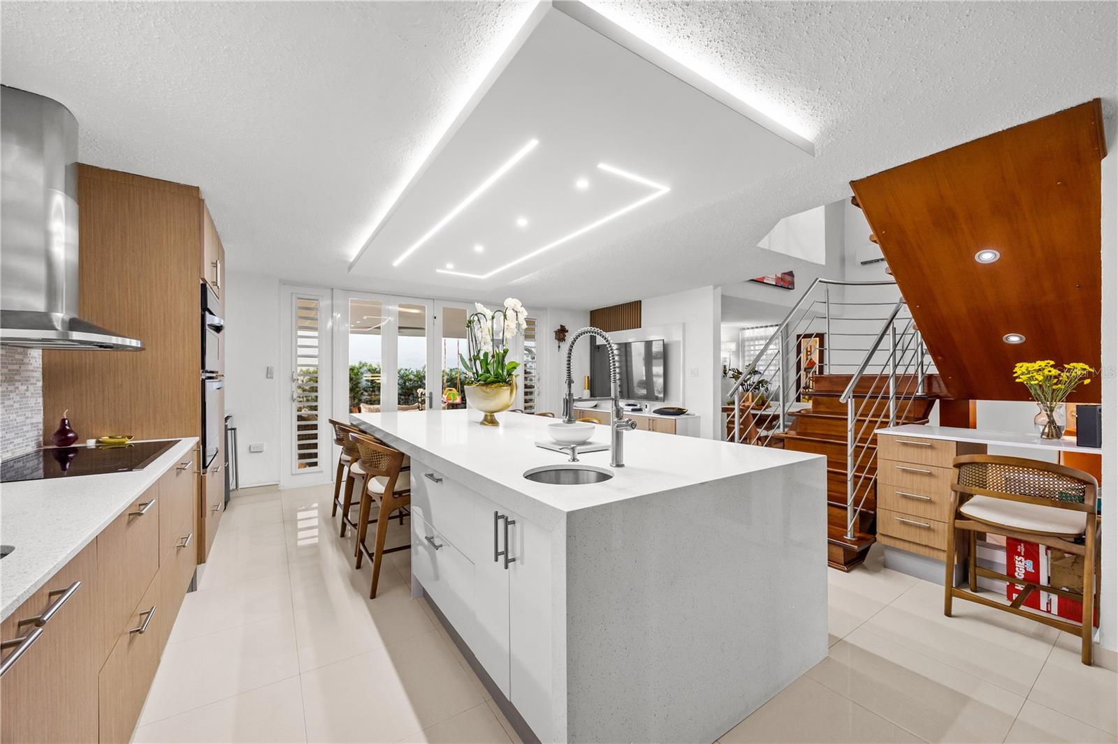 Modern kitchen with a large white island, light wood cabinets, and an open staircase.