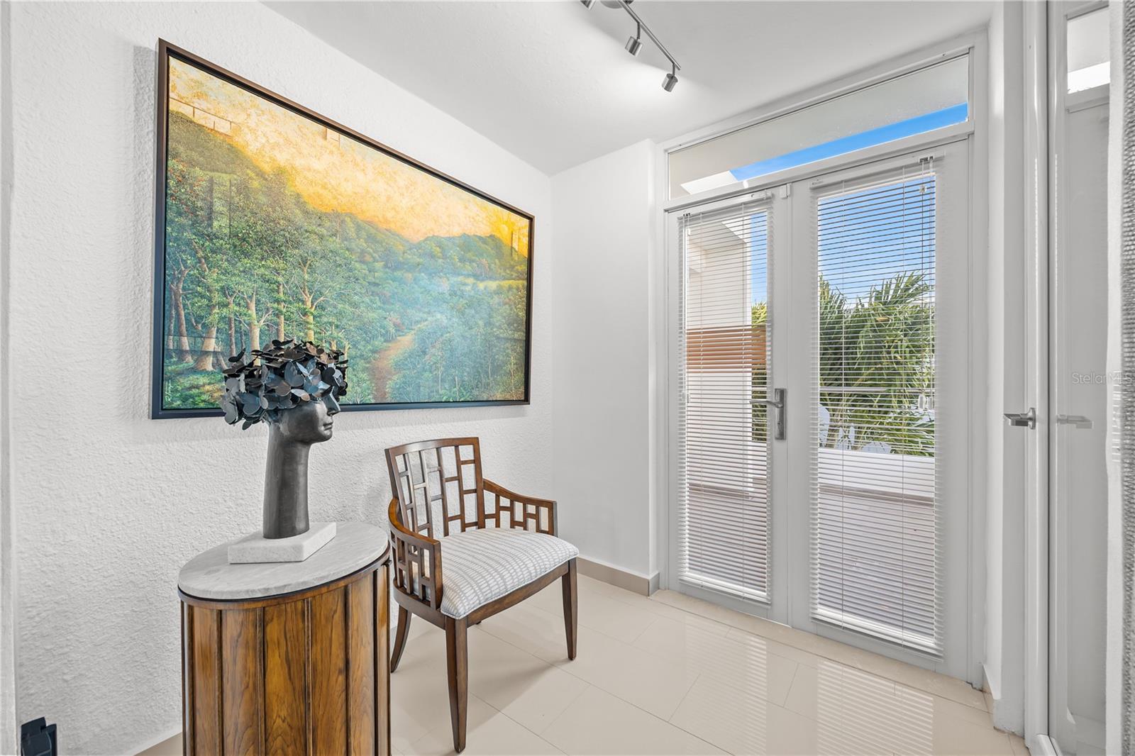 Furnished sitting area with a landscape painting, wooden chair, and glass doors overlooking tropical greenery.