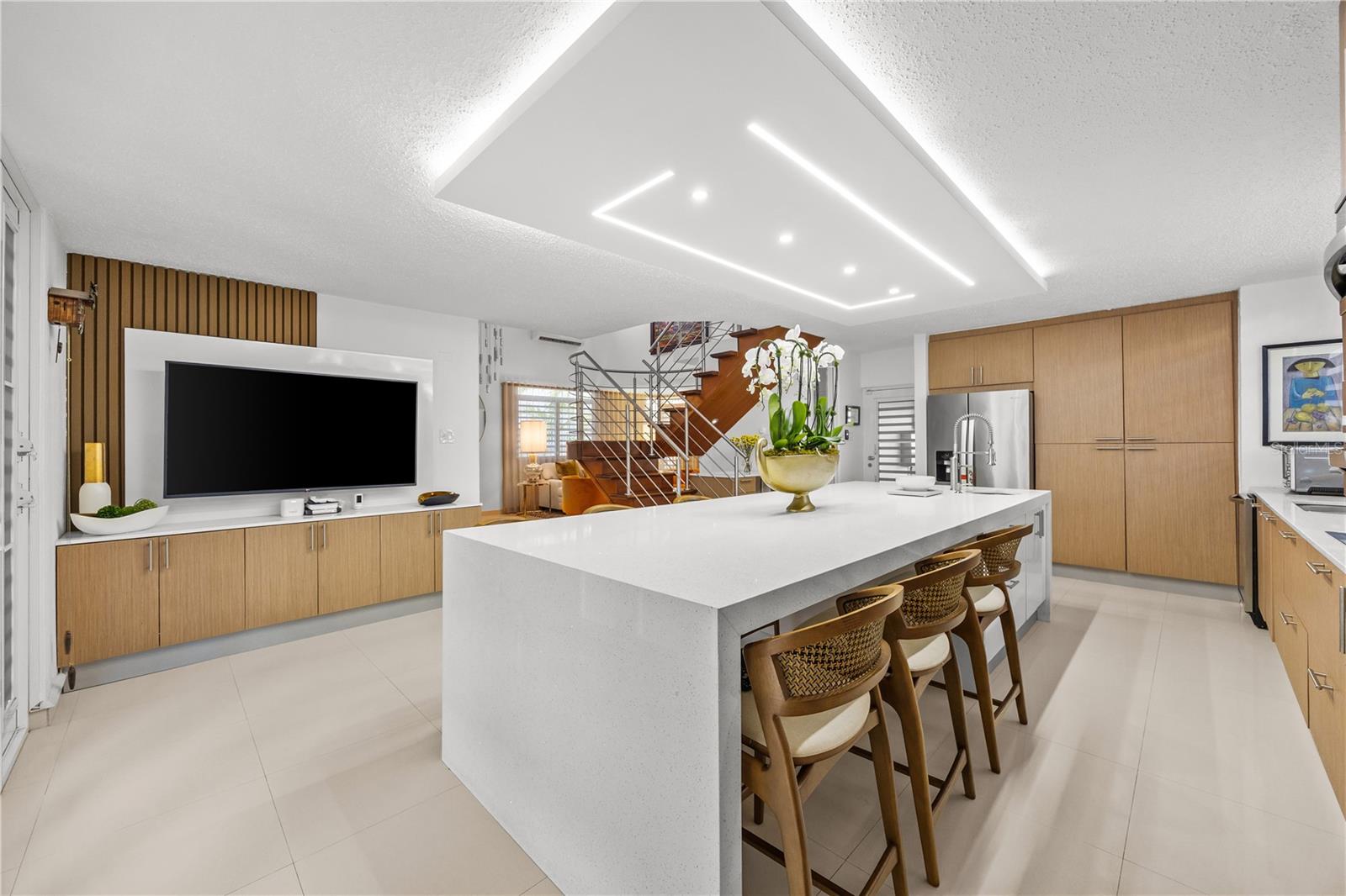 Modern kitchen with a large white island, wooden stools, and a staircase beyond.