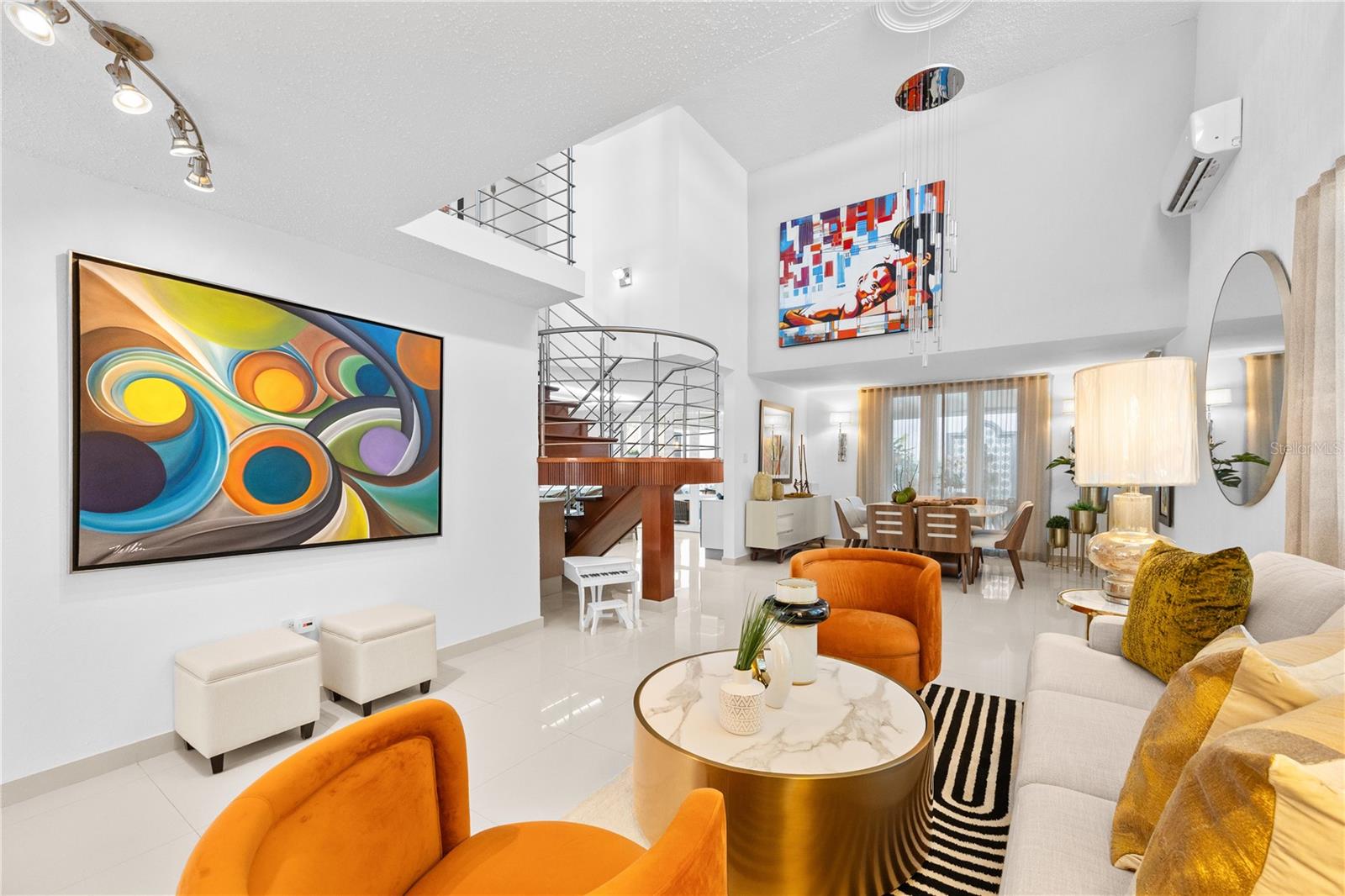 Furnished living room with colorful artwork, orange chairs, and a curved staircase.