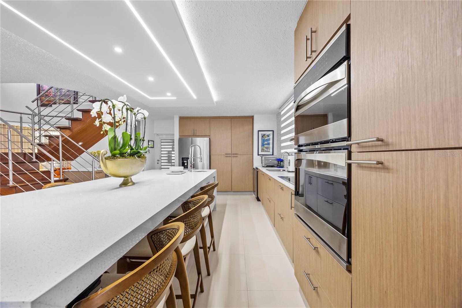 Modern kitchen with a long white island, light wood cabinets, stainless appliances, and an adjacent staircase.