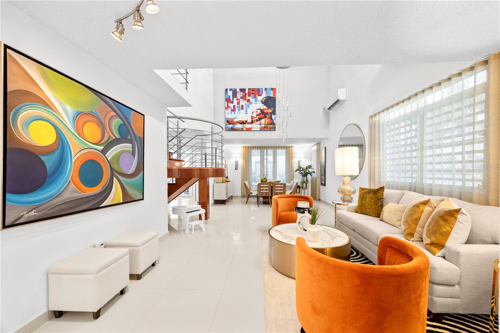 Open-plan living room with a cream sectional, vivid orange chairs, colorful art, and a curved staircase.